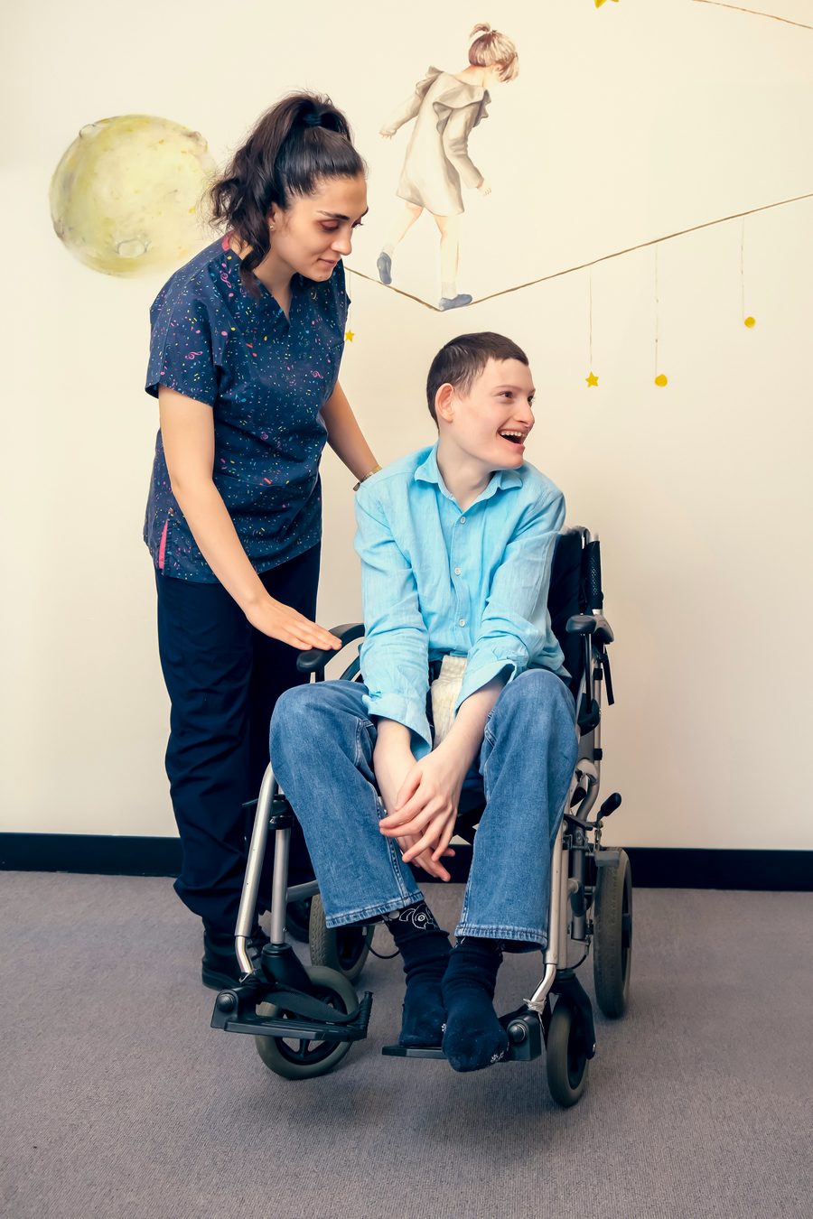 A support worker standing beside a young man in a wheelchair, both smiling.